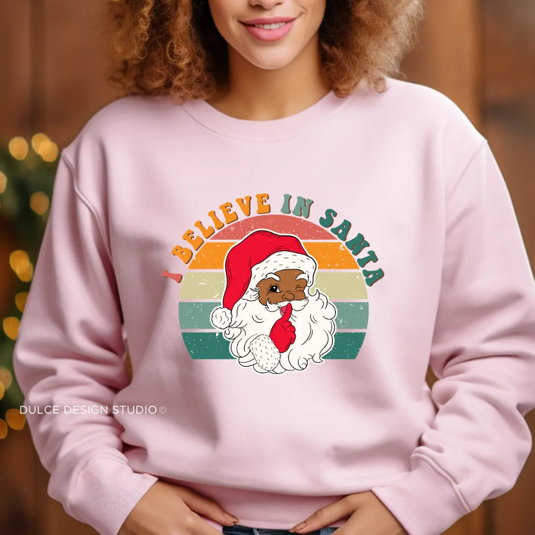 Person wearing a pink sweatshirt with a festive "I Believe in Santa" design featuring a cute Brown Skinned Santa illustration