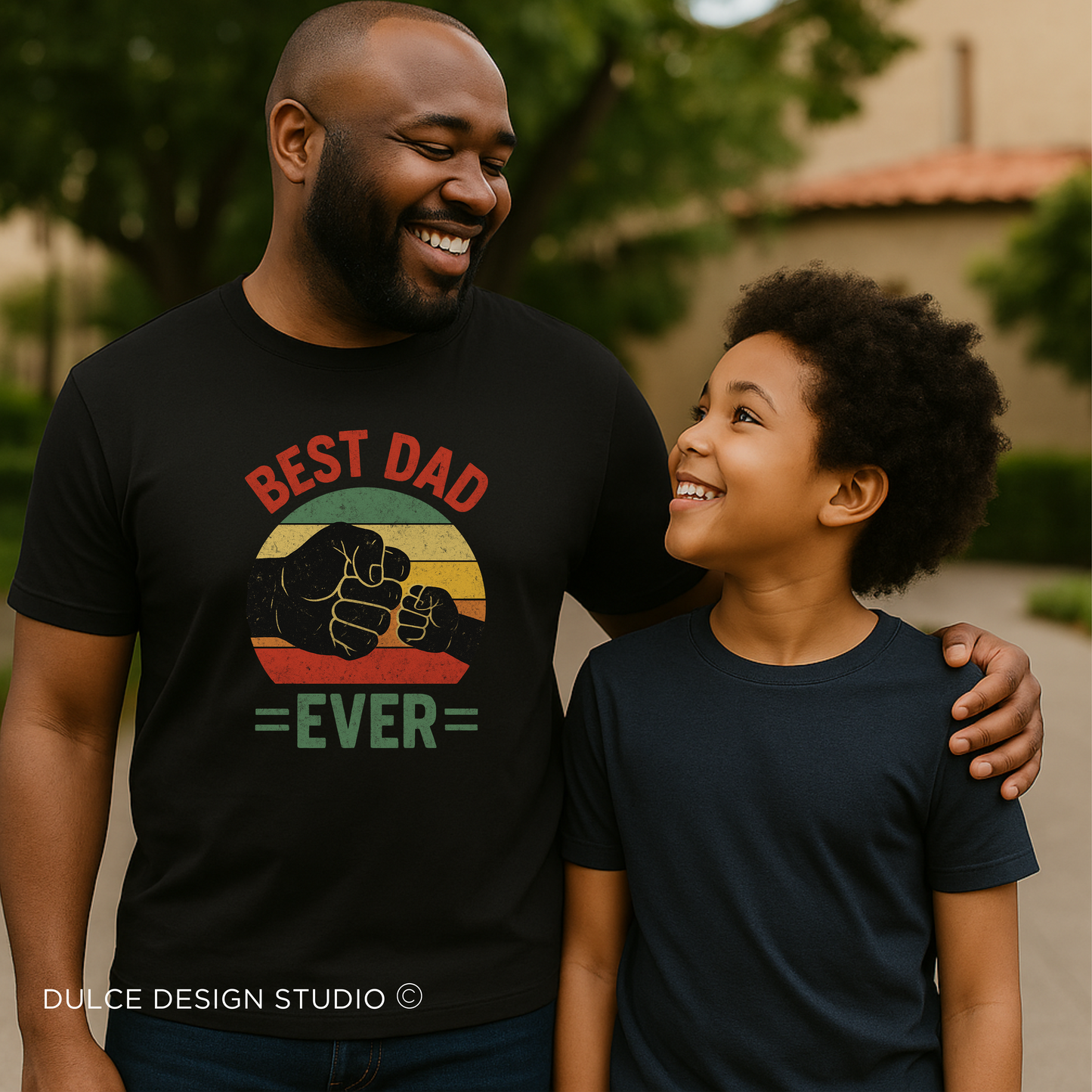 Smiling Black father and son wearing matching t-shirts; dad’s shirt features a retro-style 'Best Dad Ever' design with fist bump graphic, standing outdoors in a warm, happy moment