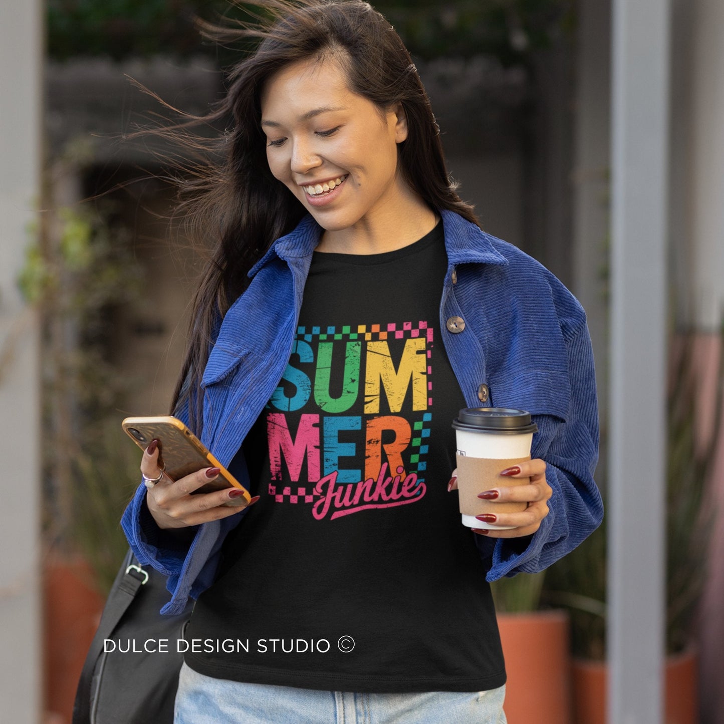 Woman wearing a black t-shirt with colorful text, holding a phone and a coffee cup,