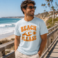 Man wearing a 'Beach Please' t-shirt by the ocean with palm trees in the background