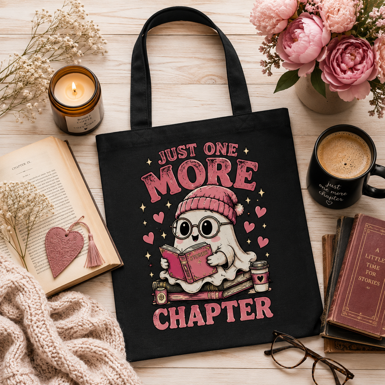 A black canvas tote bag with a cute ghost graphic reading 'JUST ONE MORE CHAPTER' in pink, styled in a cozy flat-lay with a candle, open book, flowers, mug, books, and glasses on a light wood surface.