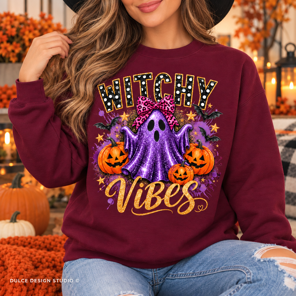 A woman wearing a burgundy crewneck sweatshirt with a colorful 'Witchy Vibes' Halloween graphic featuring a purple ghost, pumpkins, and stars, paired with blue jeans in a cozy autumn-themed setting.