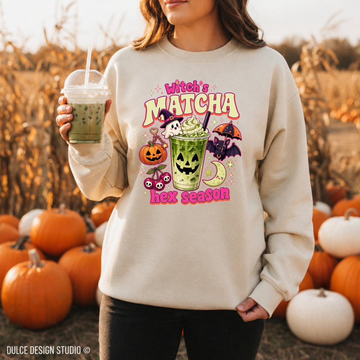 A woman wearing a cream sweatshirt with a colorful 'Witch's Matcha Hex Season' graphic, standing in a pumpkin field and holding an iced matcha drink.