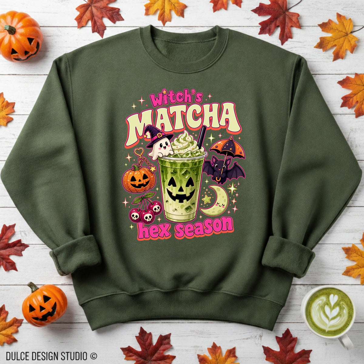 An olive green crewneck sweatshirt with a 'Witch's Matcha hex season' graphic, styled in a flat-lay on a white wood background with autumn leaves, pumpkins, and a latte.