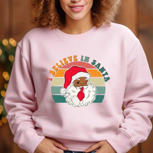 Person wearing a pink sweatshirt with a graphic of a Black Santa Claus and 'Believe in Santa' text.