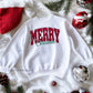 White sweatshirt with a "Merry Christmas" faux sequin design laid on a festive background with holiday decorations.