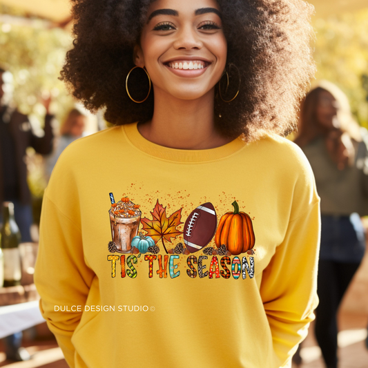 Person wearing a yellow sweatshirt with a festive design featuring a pumpkin spice latte, leaves, a football, and pumpkins with the text "Tis The Season"