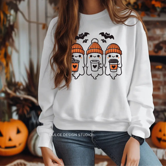 Person wearing a white sweatshirt with a design featuring three ghosts holding coffee cups, wearing orange beanies, and surrounded by bats