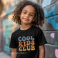little boy wearing black youth t-shirt with retro rainbow-toned text reading ‘Cool Kids Club’ in large vintage font