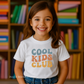little girl wearing White youth t-shirt with retro rainbow-toned text reading ‘Cool Kids Club’ in large vintage font