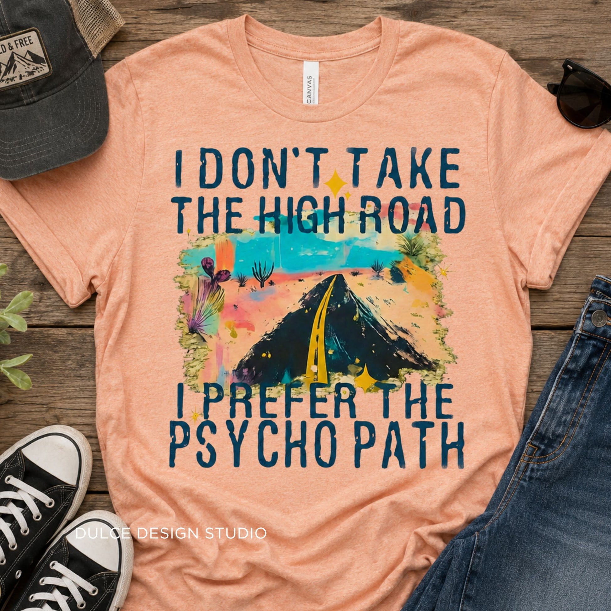 Pink t-shirt with graphic design and text on a wooden surface with shoes and a cap.
