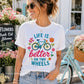Woman wearing a white t-shirt with a bicycle design and 'Life is better on two wheels' text, standing next to a bicycle in a floral setting.
