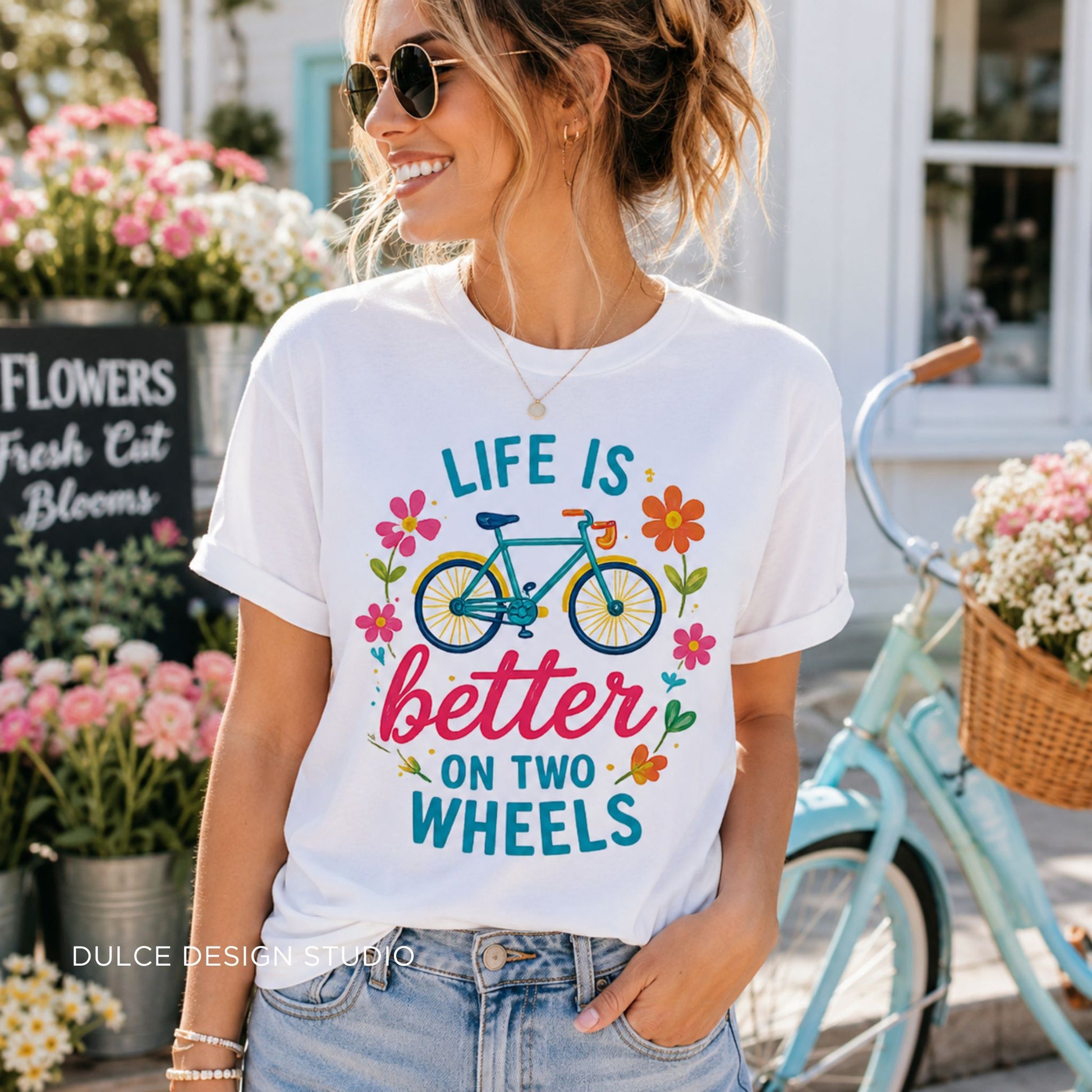 Woman wearing a white t-shirt with a bicycle design and 'Life is better on two wheels' text, standing next to a bicycle in a floral setting.