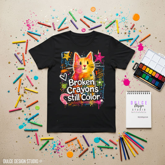 Black t-shirt with a colorful graphic of a cat and 'Broken Crayons Still Color' text, surrounded by crayons and art supplies on a beige background.