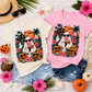 Two graphic t-shirts, one cream and one pink, featuring a cartoon ghost with a pink nose in a Halloween tropical setting, styled in a flat lay with denim shorts and autumn props.