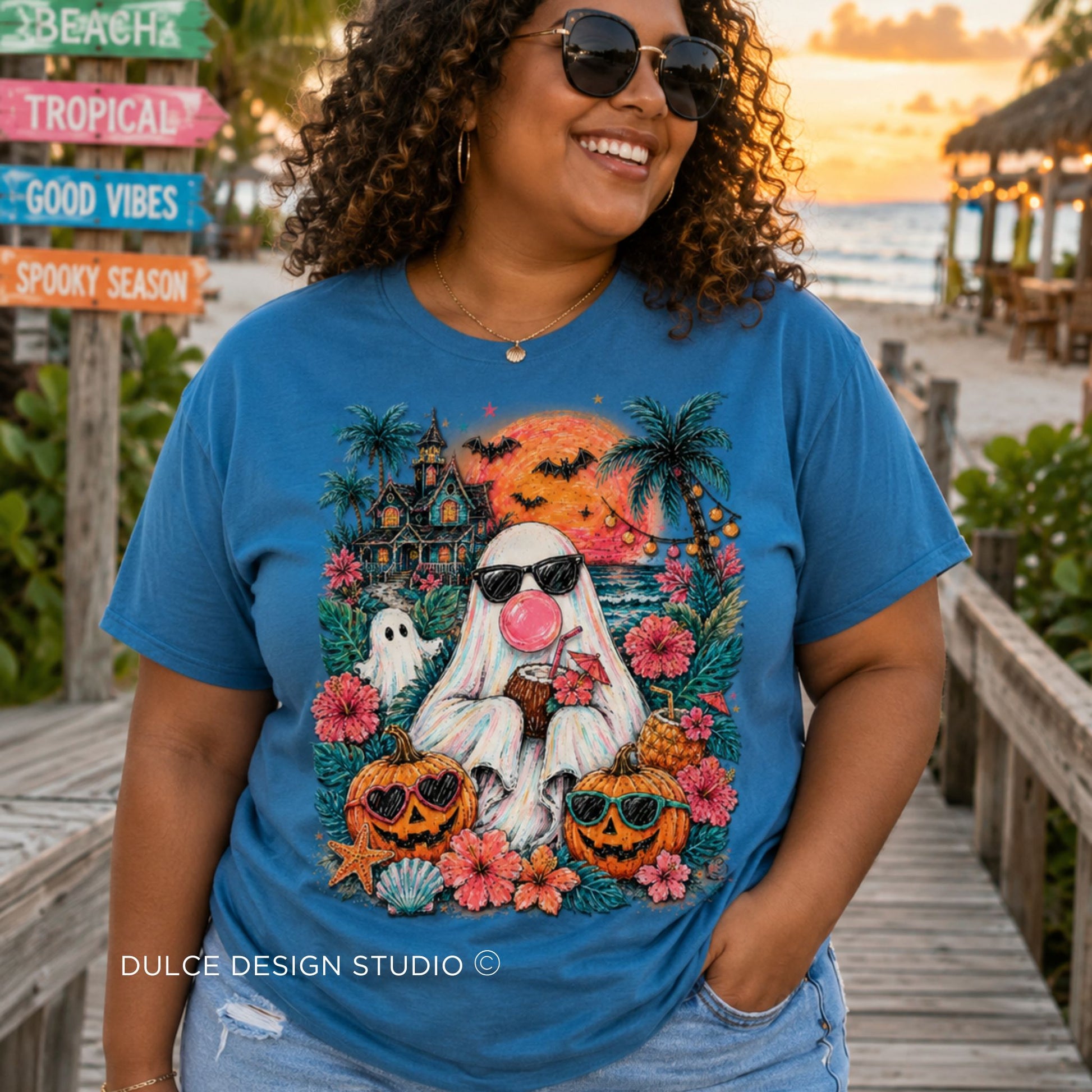 A woman wearing a blue graphic t-shirt with a Halloween-themed design featuring a ghost, pumpkins, flowers, and a sunset beach background. The t-shirt is branded with 'DULCE DESIGN STUDIO' text at the bottom.