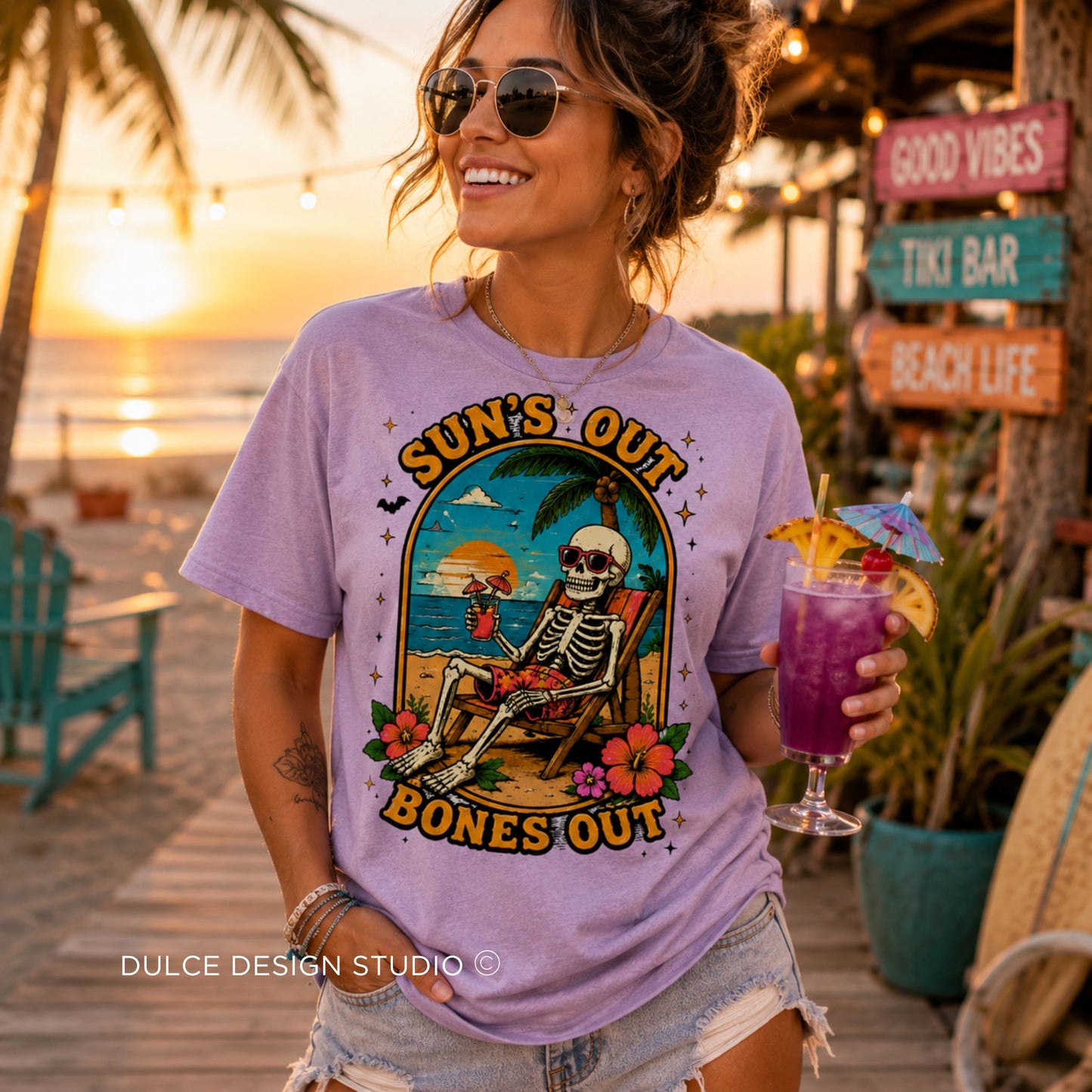 A woman wearing a lavender graphic t-shirt with a skeleton beach design and the text 'SUN'S OUT BONES OUT', holding a purple cocktail on a beach patio at sunset.