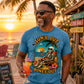 A man wearing a blue graphic t-shirt with a skeleton beach design and the text 'SUN'S OUT BONES OUT' stands on a beach at sunset holding a cocktail. The watermark 'DULCE DESIGN STUDIO' is visible at the bottom.