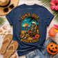 A navy blue Bella Canvas graphic t-shirt featuring a skeleton sitting in a beach chair with the text 'SUN'S OUT BONES OUT' in a retro sunset design. The shirt is styled in a flat lay with a straw hat, sunglasses, flip-flops, denim shorts, a jack-o'-lantern, and tropical flowers on a white wood background.