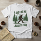 White t-shirt with a graphic design featuring a bigfoot and text on a wooden surface with pinecones and a map.