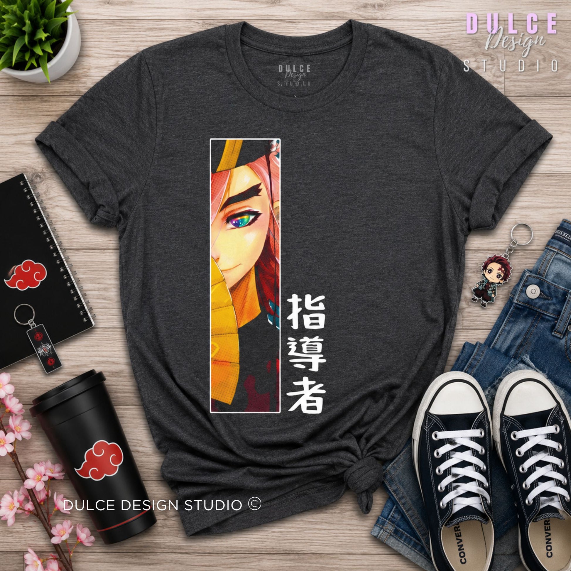 A dark heather gray graphic t-shirt featuring an anime character print and Japanese text, styled in a flat-lay arrangement on a wooden surface with jeans, sneakers, a coffee cup, and a notebook.