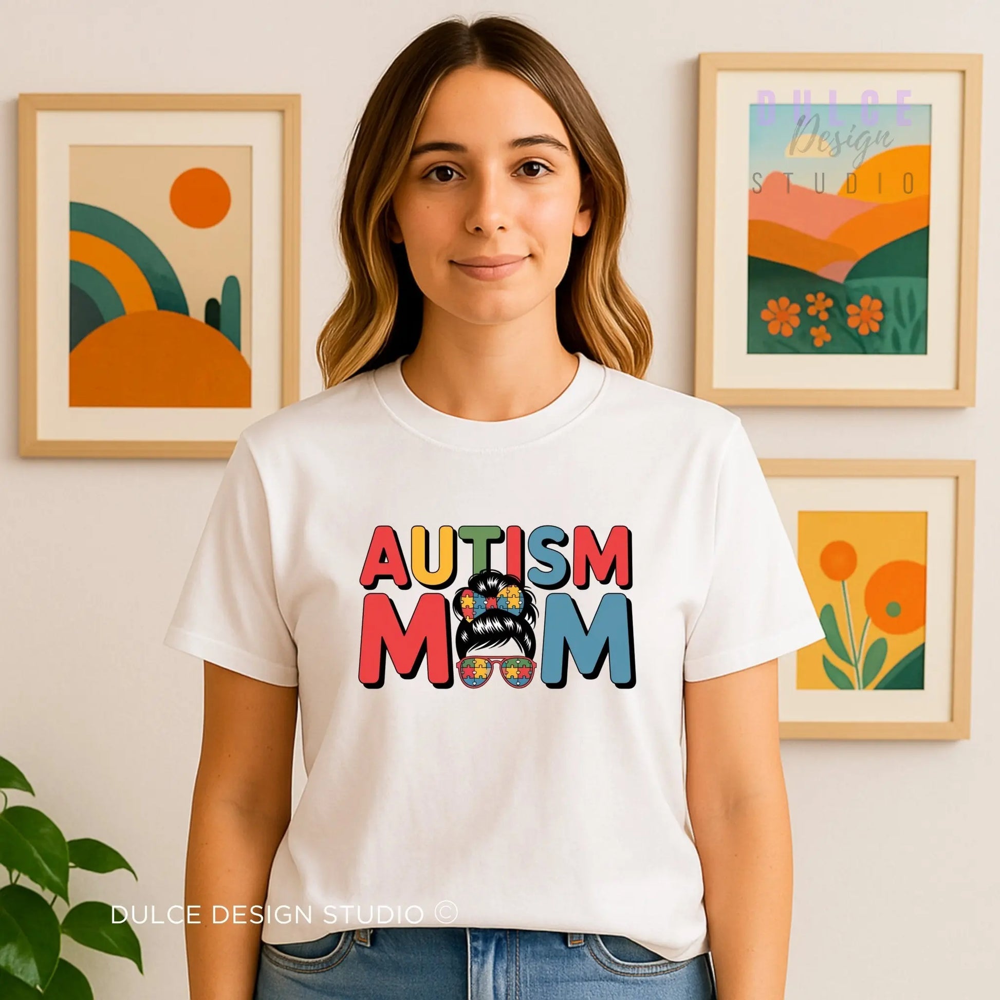 Woman wearing a white t-shirt with 'AUTISM MOM' text in colorful letters, standing in front of wall art.