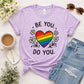 Purple t-shirt with rainbow heart and text 'Be You, Do You' on a white surface with accessories.