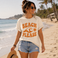 Woman on a beach wearing a 'Beach Please' t-shirt with sunglasses and a hat.
