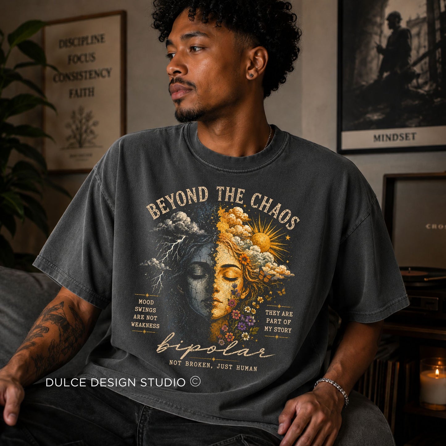 Man wearing a t-shirt with a graphic design and text, sitting in a room with decor elements.