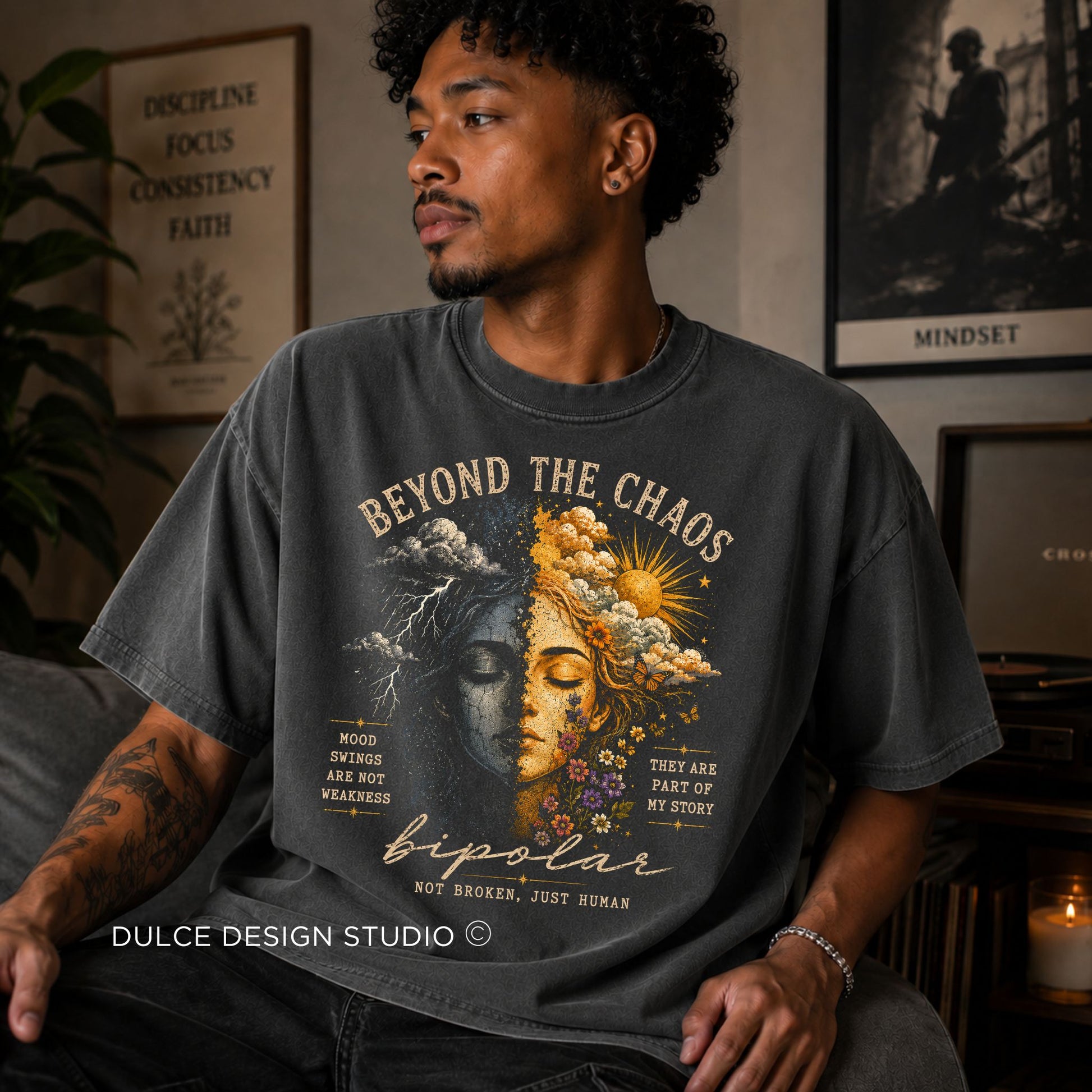 Man wearing a t-shirt with a graphic design and text, sitting in a room with decor elements.