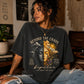 Person wearing a black t-shirt with a graphic design and text, sitting in a room with decor.