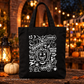 A black canvas tote bag with a white hand-drawn Halloween graphic, displayed on a rustic wooden table with a lit lantern, a small pumpkin, and a warm, blurred outdoor background.
