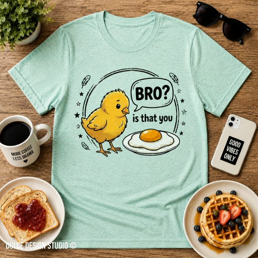 Light blue t-shirt with a cartoon chicken and egg design on a wooden table with breakfast items.