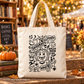 A cream canvas tote bag with black hand-drawn Halloween graphics, including a jack-o'-lantern, ghosts, bats, spiders, and spooky text, standing on a wooden table in a warm, blurred outdoor market setting with a pumpkin and books nearby.