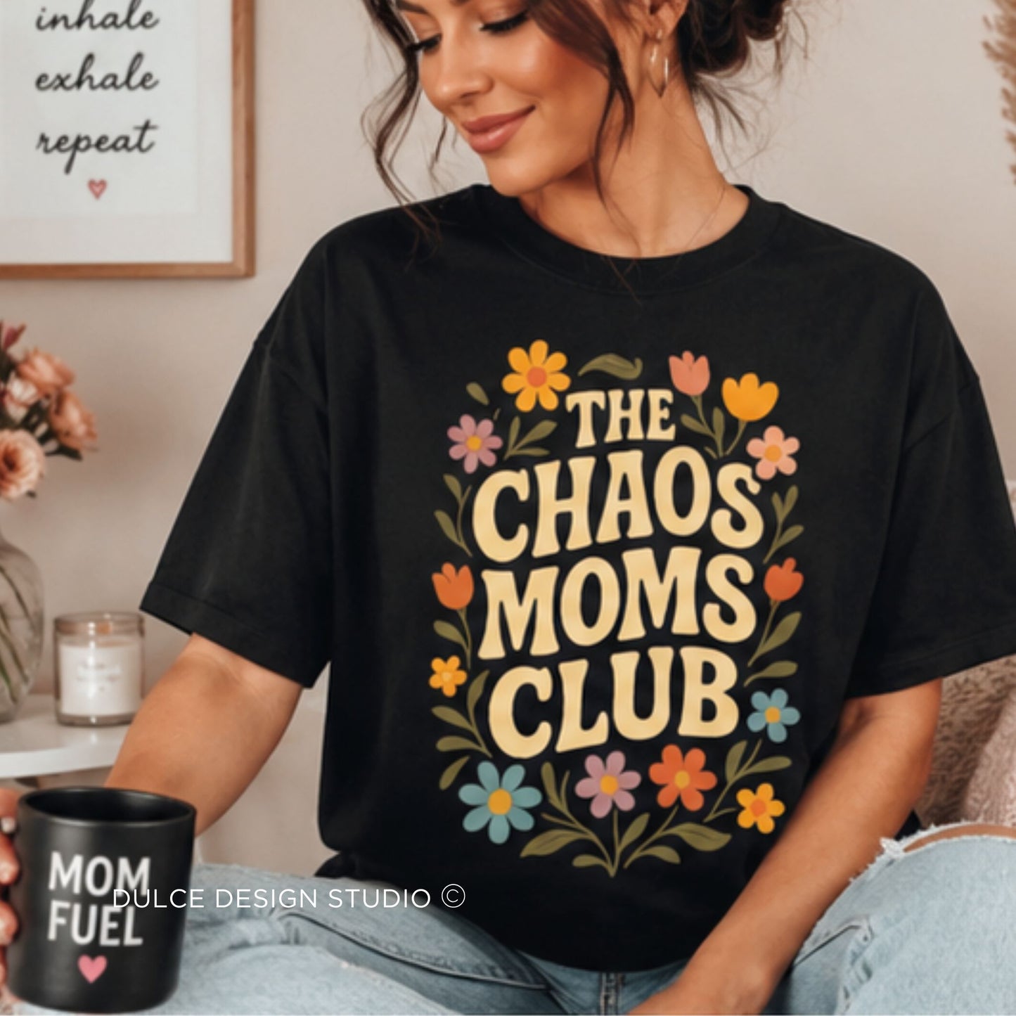 A woman wearing a black graphic t-shirt with a retro floral design that reads 'THE CHAOS MOMS CLUB' in beige letters, surrounded by colorful flowers. She is holding a black mug with 'MOM FUEL' text.