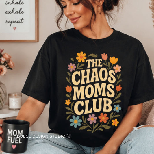 A woman wearing a black graphic t-shirt with a retro floral design that reads 'THE CHAOS MOMS CLUB' in beige letters, surrounded by colorful flowers. She is holding a black mug with 'MOM FUEL' text.