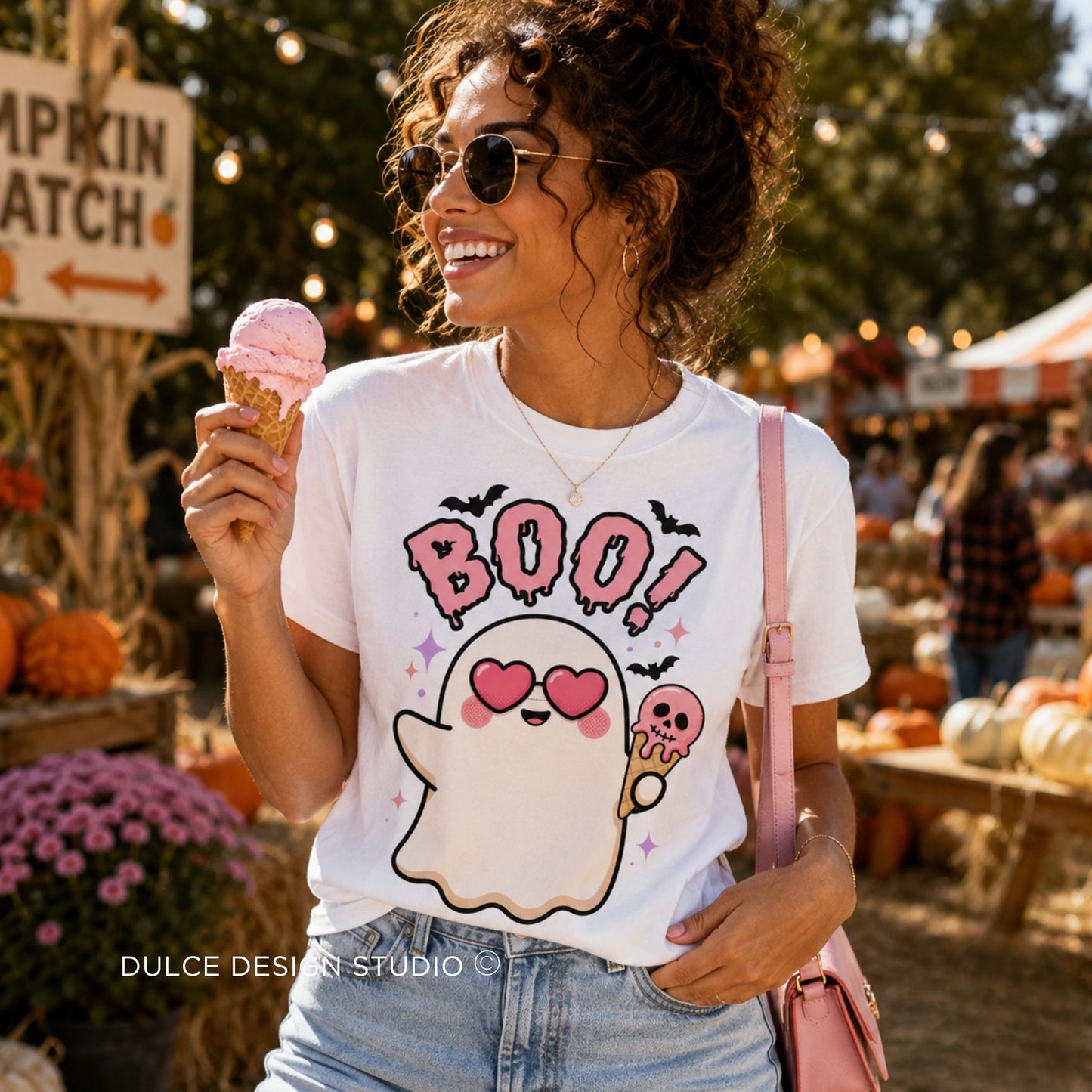 A woman at an outdoor autumn market wearing a white graphic t-shirt with a cute ghost holding a pink ice cream cone and the word 'BOO!' in pink. She is holding a pink ice cream cone and wearing a pink shoulder bag.