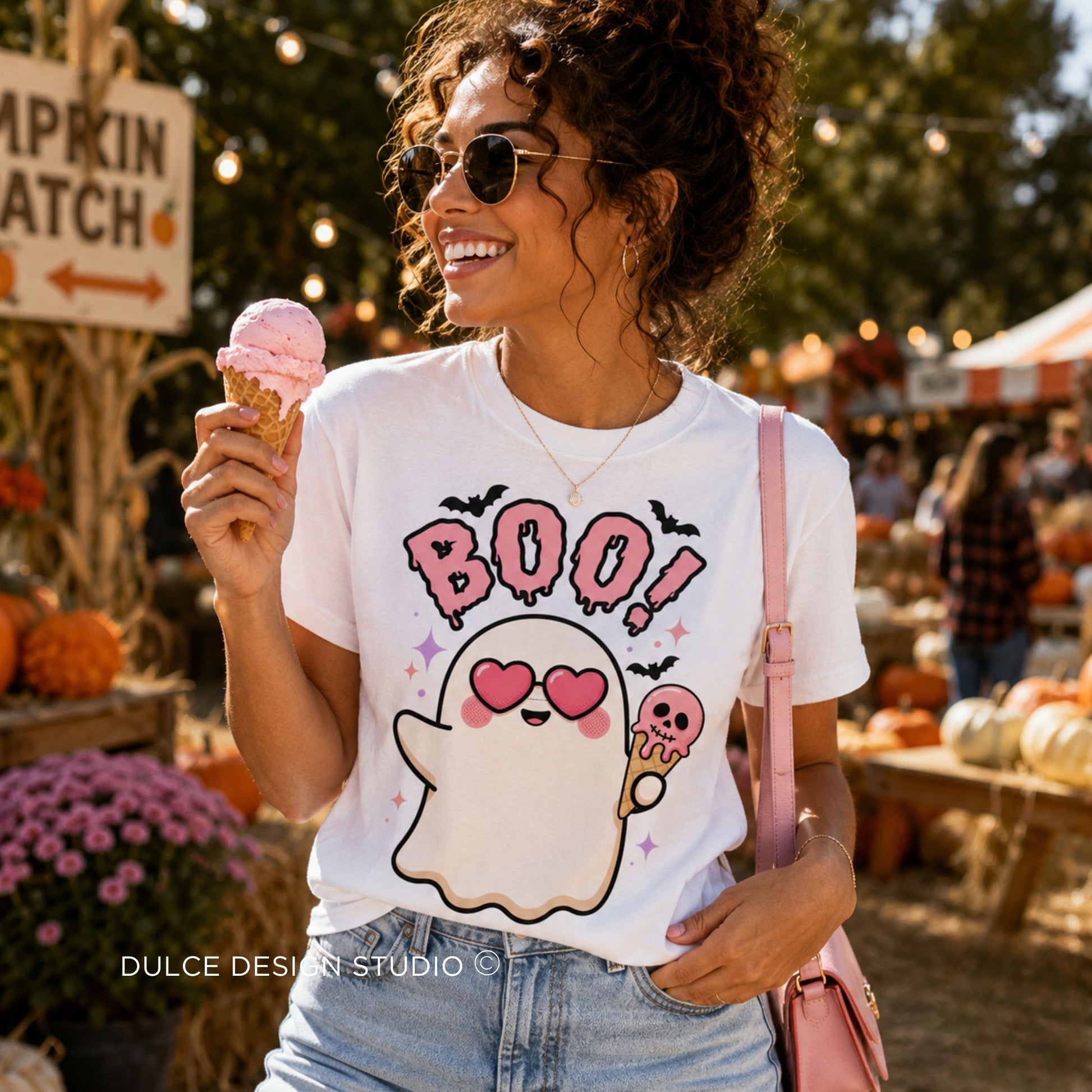 A woman at an outdoor autumn market wearing a white graphic t-shirt with a cute ghost holding a pink ice cream cone and the word 'BOO!' in pink. She is holding a pink ice cream cone and wearing a pink shoulder bag.