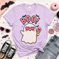 A light purple short-sleeve graphic T-shirt laid flat on a white wood surface, styled with Halloween props including sunglasses, a small pumpkin, candy, lollipops, jeans, and black shoes. The shirt has a large cartoon ghost with pink heart eyes, pink 'BOO!' text, and small bat/star accents.