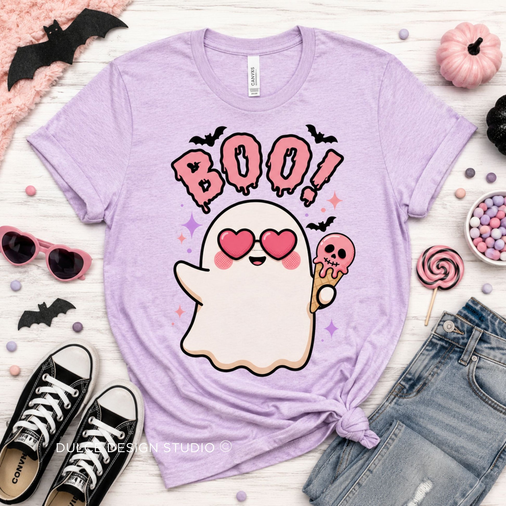 A light purple short-sleeve graphic T-shirt laid flat on a white wood surface, styled with Halloween props including sunglasses, a small pumpkin, candy, lollipops, jeans, and black shoes. The shirt has a large cartoon ghost with pink heart eyes, pink 'BOO!' text, and small bat/star accents.