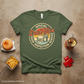 military Green t-shirt with 'Official Dad Tax Food Inspection Service' graphic on a textured surface with food items.