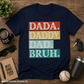 Navy blue t-shirt with colorful text 'DADA. DADDY. DAD. BRUH.' on a wooden surface with accessories.