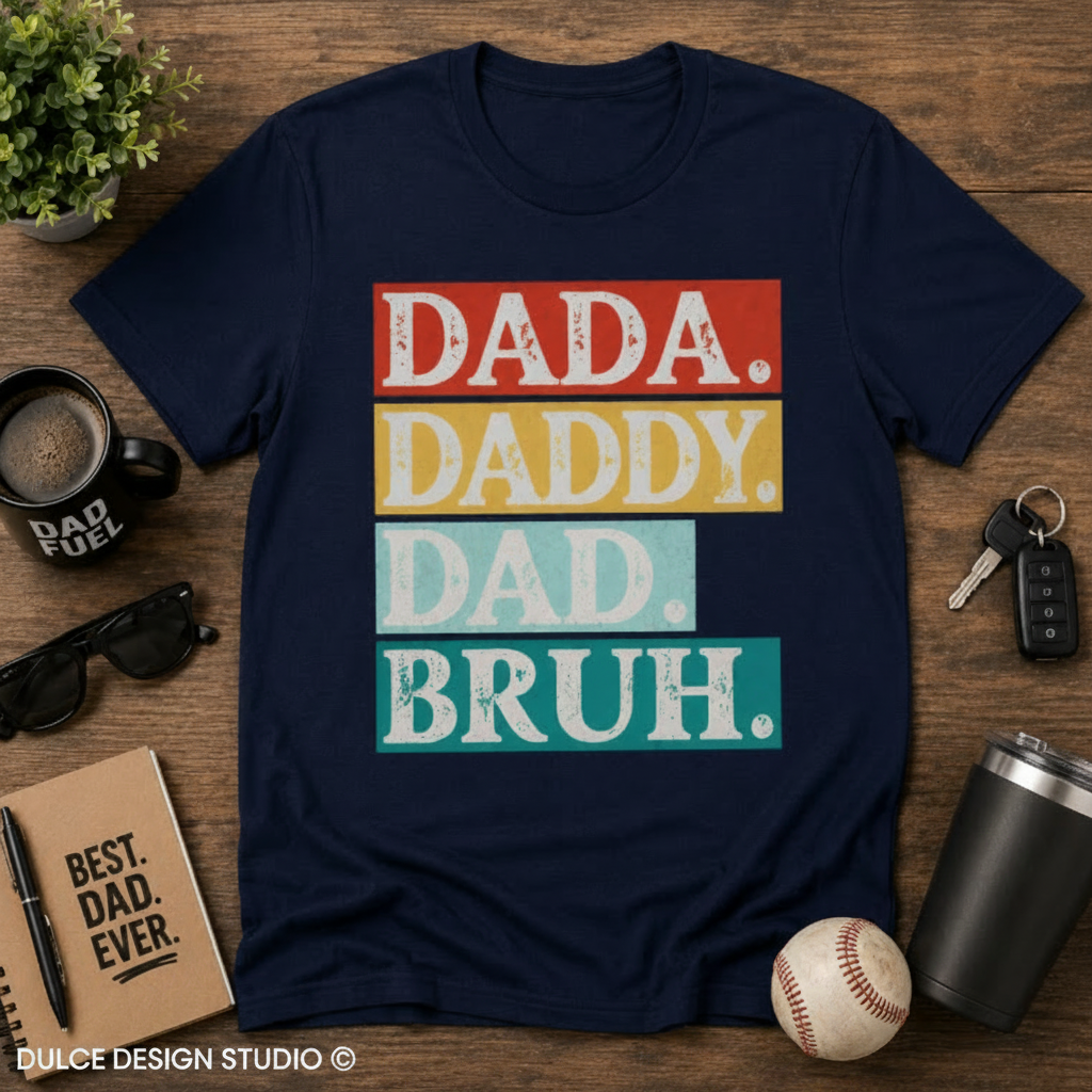 Navy blue t-shirt with colorful text 'DADA. DADDY. DAD. BRUH.' on a wooden surface with accessories.