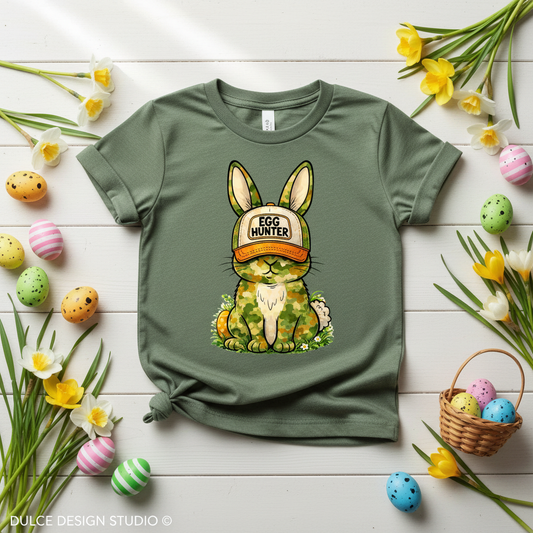 Green t-shirt with a bunny wearing an 'Egg Hunter' cap on a wooden surface with Easter decorations.
