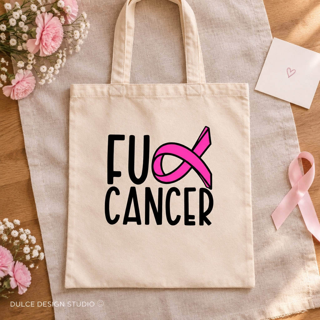 A beige canvas tote bag laid flat on a linen cloth over a wooden surface, printed with the words 'FU CANCER' in black and a bright pink ribbon graphic. Pink flowers, a small card, and a pink ribbon are arranged around it.