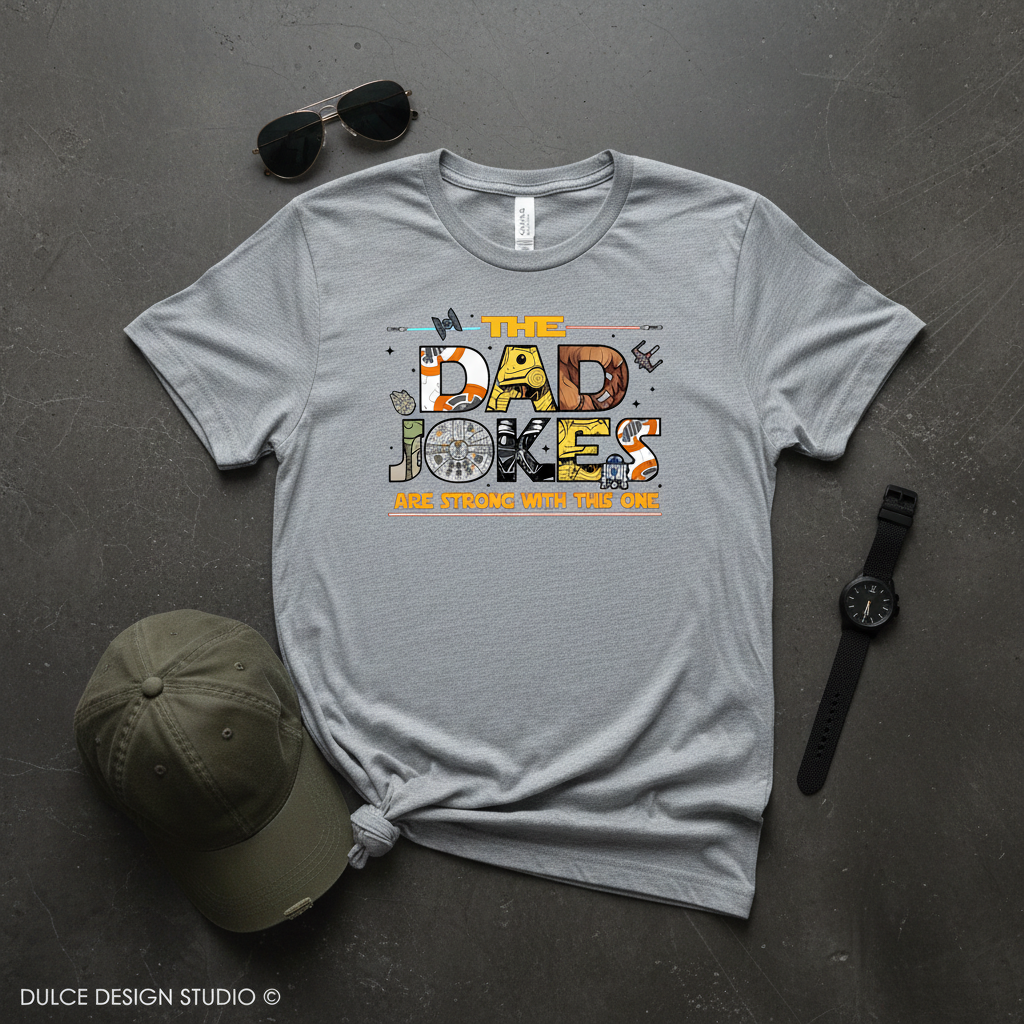 Gray t-shirt with 'The Dad Jokes Are Strong With This One' design on a dark surface with a cap, sunglasses, and watch.