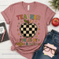 A heather mauve short-sleeve graphic T-shirt laid flat on a white wood surface, styled with a notebook, pink pen, red apple, blue jeans, glasses, and sticky notes. The shirt graphic reads “TEACHER FUELED BY coffee & patience” with a checkerboard apple in the center.