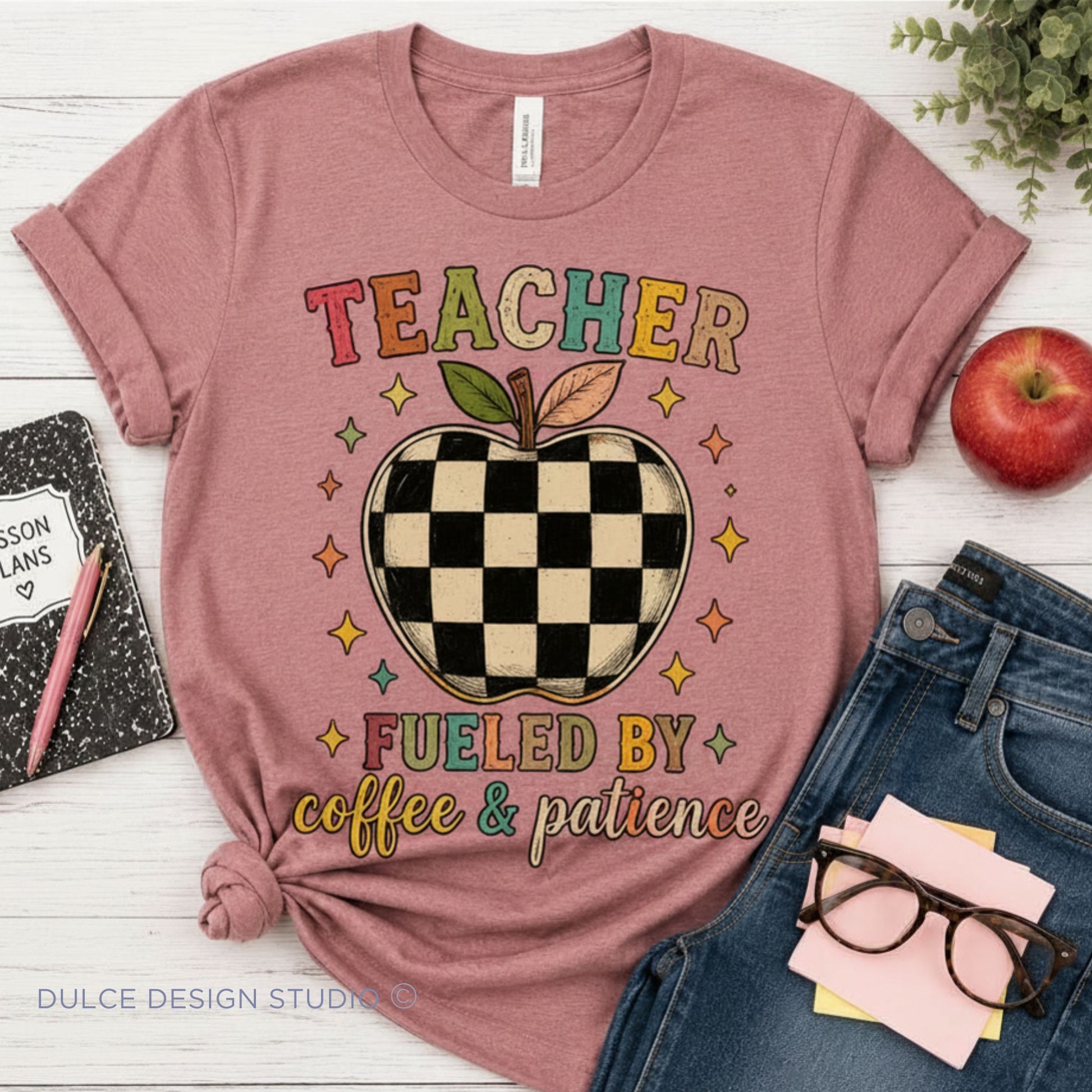 A heather mauve short-sleeve graphic T-shirt laid flat on a white wood surface, styled with a notebook, pink pen, red apple, blue jeans, glasses, and sticky notes. The shirt graphic reads “TEACHER FUELED BY coffee & patience” with a checkerboard apple in the center.