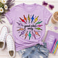 A styled flat-lay featuring a light purple t-shirt with a colorful lightning-bolt graphic and the text 'good vibes only'. The t-shirt is styled with denim shorts, white Converse sneakers, a purple notebook, sunglasses, and flowers on a white wood background.
