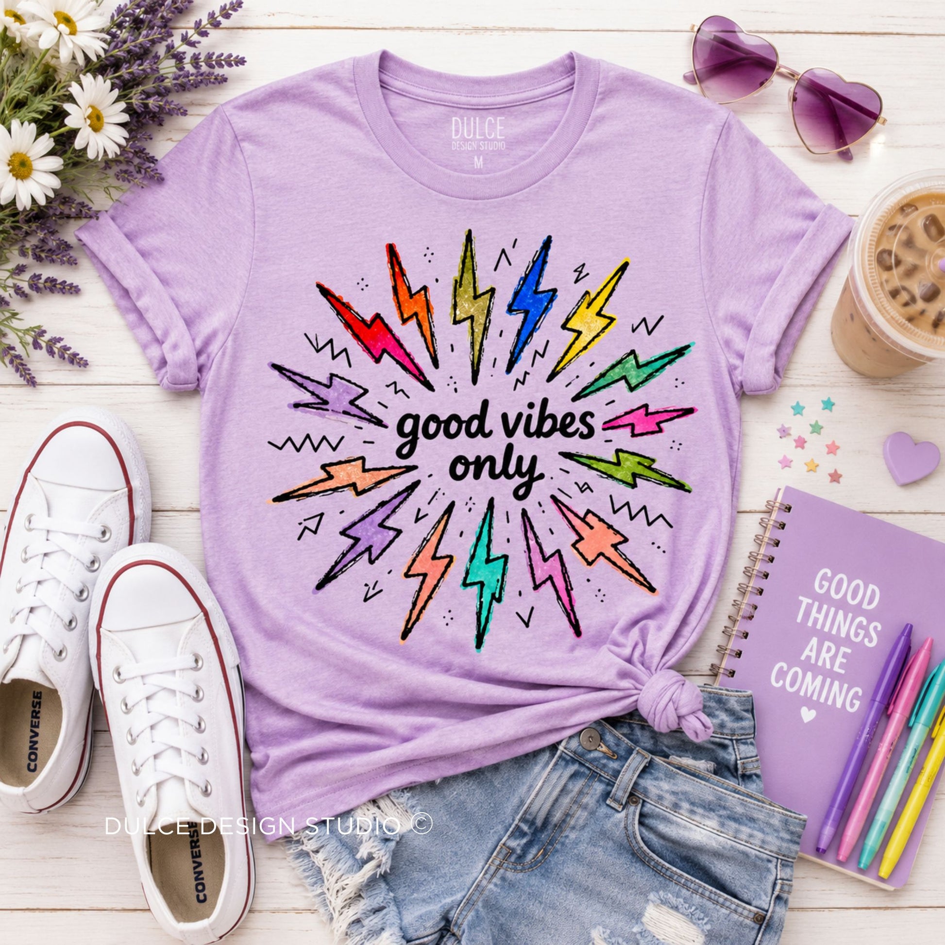 A styled flat-lay featuring a light purple t-shirt with a colorful lightning-bolt graphic and the text 'good vibes only'. The t-shirt is styled with denim shorts, white Converse sneakers, a purple notebook, sunglasses, and flowers on a white wood background.